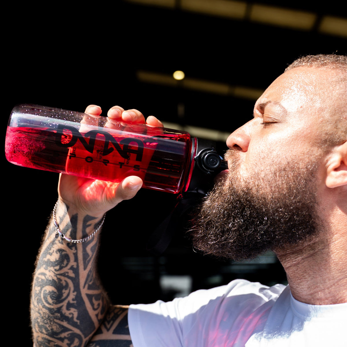 A man with a beard and tattoos drinking from a red transparent bottle with a black cap, wearing a white t-shirt with 'MADE OF MORE' and 'Dna Sports' printed on it, set against a dark background.
